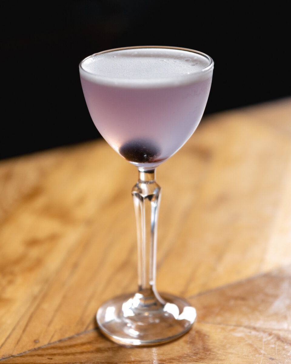 A pale purple cocktail in a stemmed glass with a single dark cherry at the bottom.