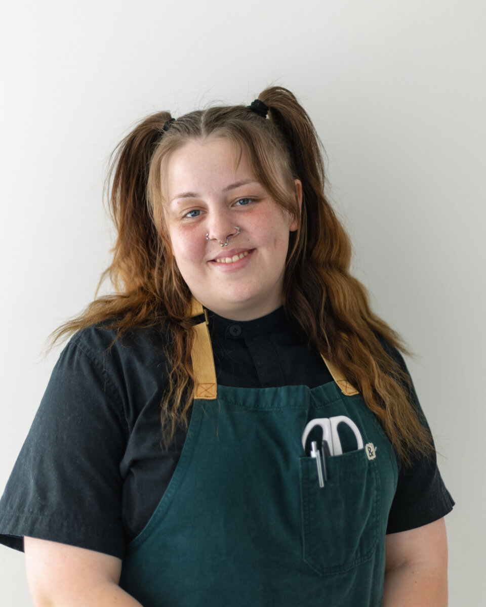 A smiling person wears a black shirt and a green apron with tools in the pocket, ready to share the food history of North of Bourbon.