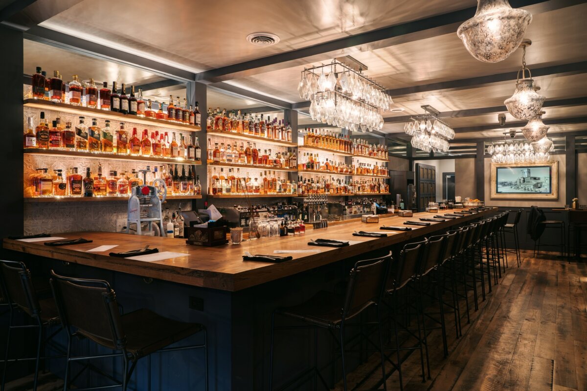 A long wooden bar with stools and illuminated shelves stocked with numerous liquor bottles.