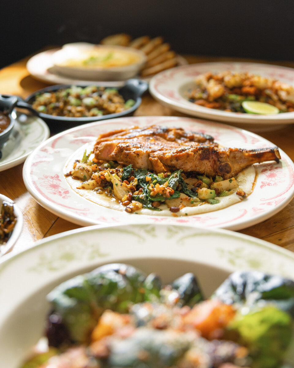 A plate featuring a grilled bone-in pork chop served over mashed potatoes and sautéed greens, surrounded by other dishes on a wooden table.