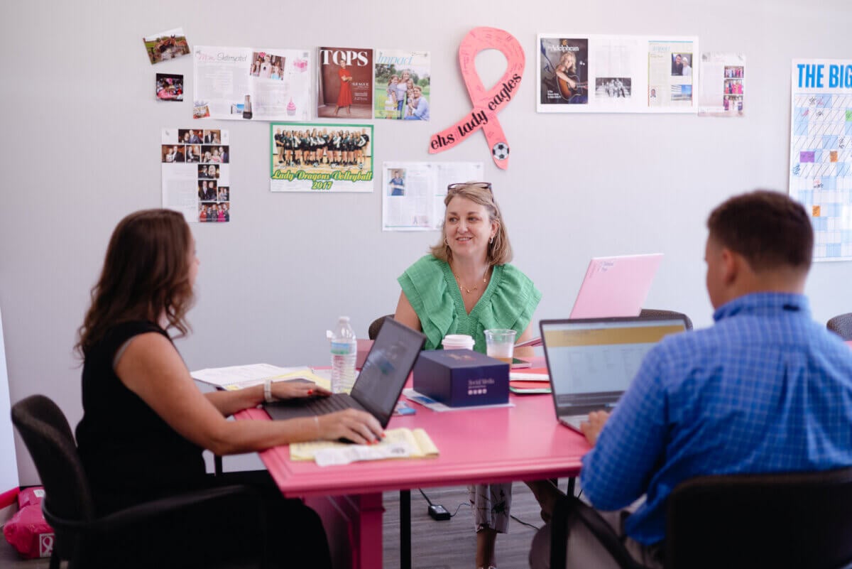 She Turned Her Darkest Moment Into a $2M Research Legacy. And She's Not Done Yet - 4 Three people sit at a pink table with laptops and papers in an office, beneath photos, a calendar, and a large pink ribbon on the wall.