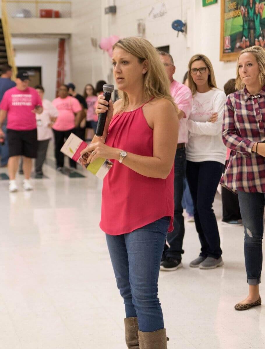 She Turned Her Darkest Moment Into a $2M Research Legacy. And She's Not Done Yet - 3 A woman in a pink top speaks into a microphone about her research legacy while holding papers, standing indoors with a group of people behind her.