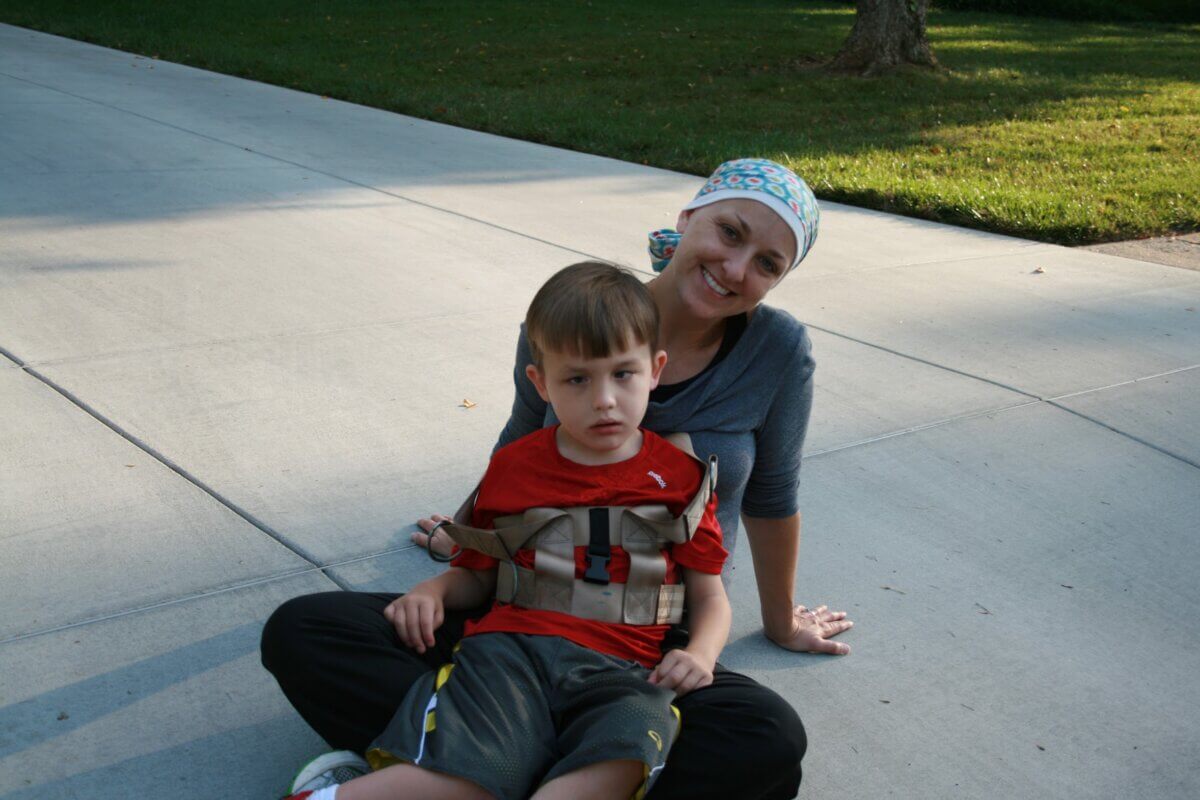 She Turned Her Darkest Moment Into a $2M Research Legacy. And She's Not Done Yet - 6 A woman sits on a sidewalk with a young boy in a red shirt wearing a harness, finding hope even in their darkest moment. Grass and a tree are visible in the background.