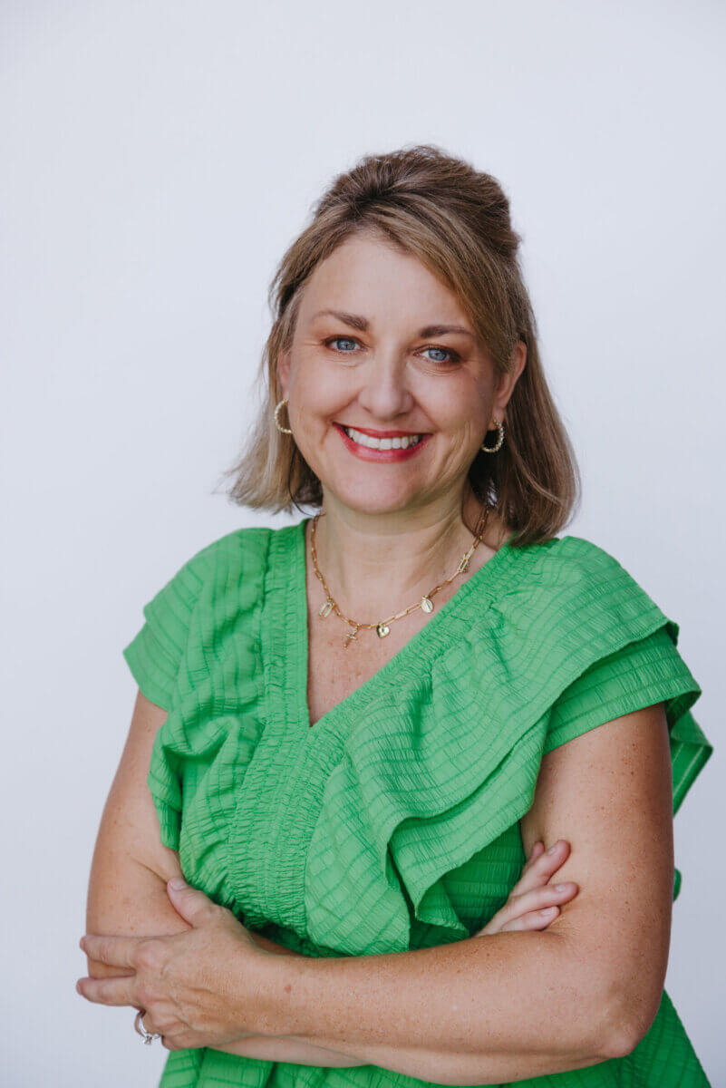 She Turned Her Darkest Moment Into a $2M Research Legacy. And She's Not Done Yet - 2 Woman with shoulder-length light brown hair and a green dress stands against a plain white background, smiling with arms crossed.