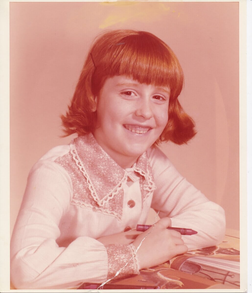 Smiling child with short brown hair sits at a desk holding a crayon, wearing a white shirt with a decorative collar, with books and a coloring page in front of her.