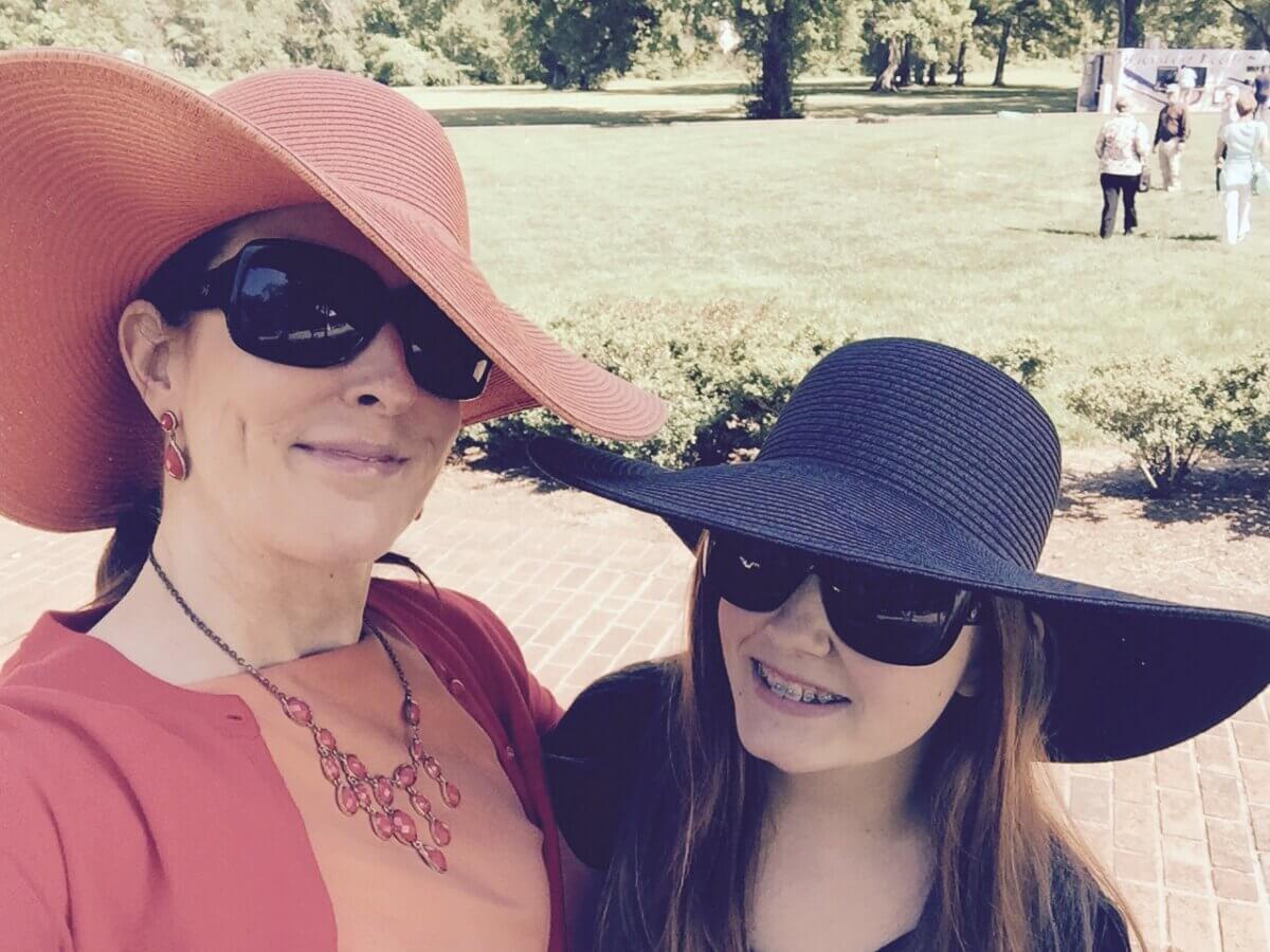 Two people wearing large sun hats and sunglasses smile for a selfie outdoors on a sunny day, with grass and trees in the background.