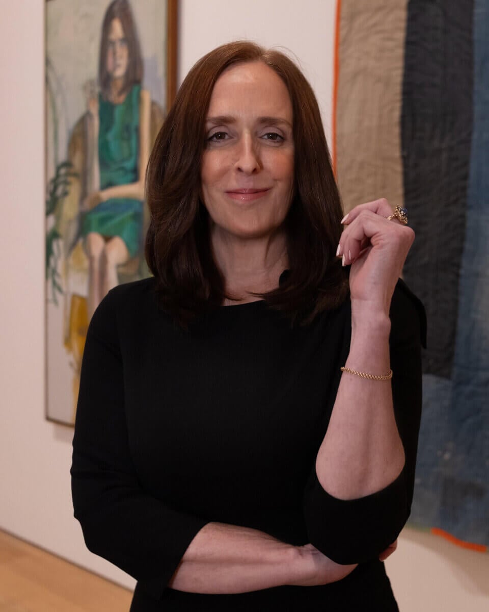 Kathleen Jameson, with shoulder-length brown hair in a black dress, stands confidently with arms crossed before a painting of a seated figure in an art gallery.