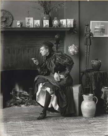 Woman in Victorian-era clothing sits by a fireplace, holding a teacup and cigarette, surrounded by pottery, framed photos, and decorative items on the mantel.