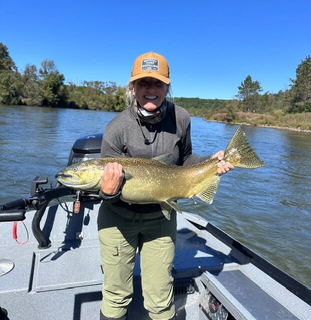 A woman in a baseball cap stands on a boat holding a large salmon.
