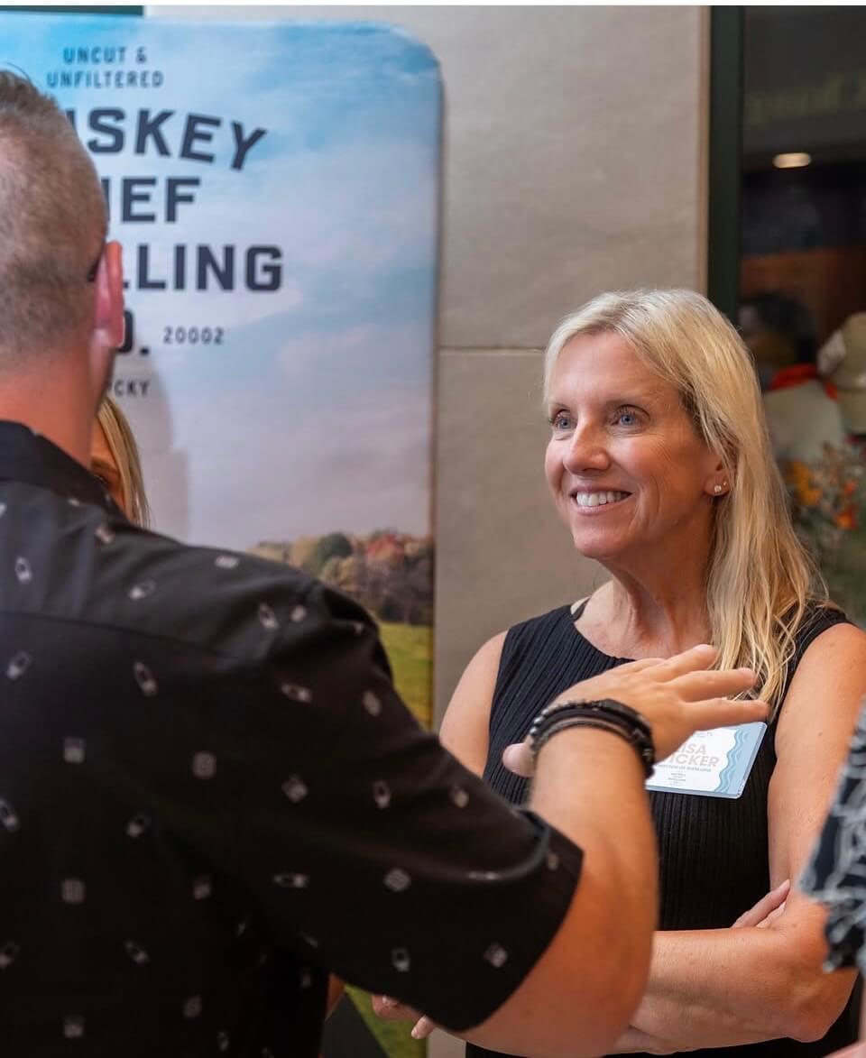 A woman with blonde hair smiling while speaking to a man at an event.