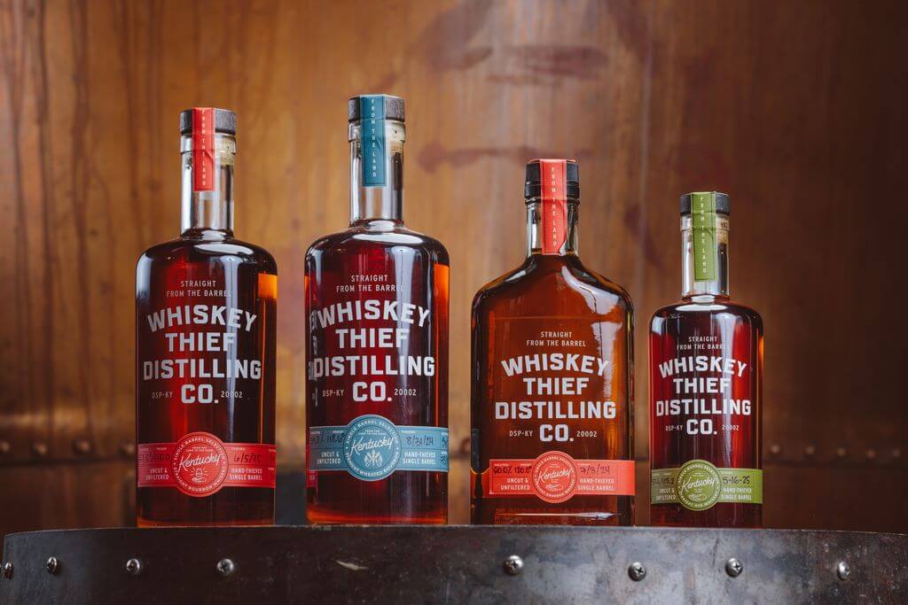Four bottles of Whiskey Thief Distilling Co. bourbon, arranged in a row on a wooden barrel.