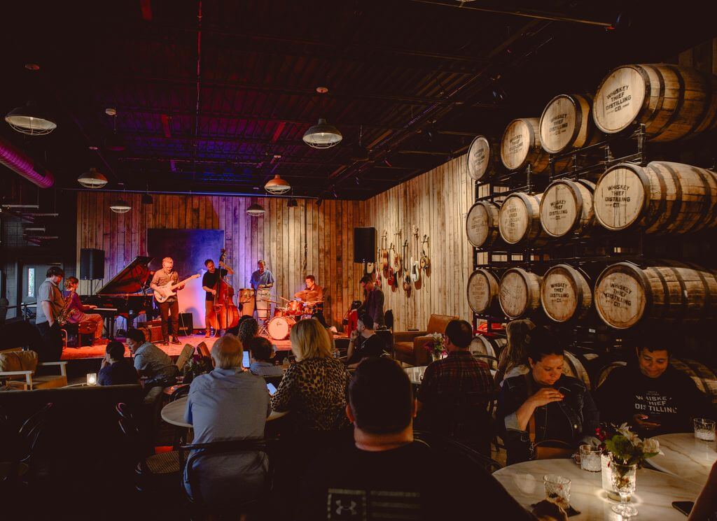 A band performs on a stage in a dim bar lined with whiskey barrels.