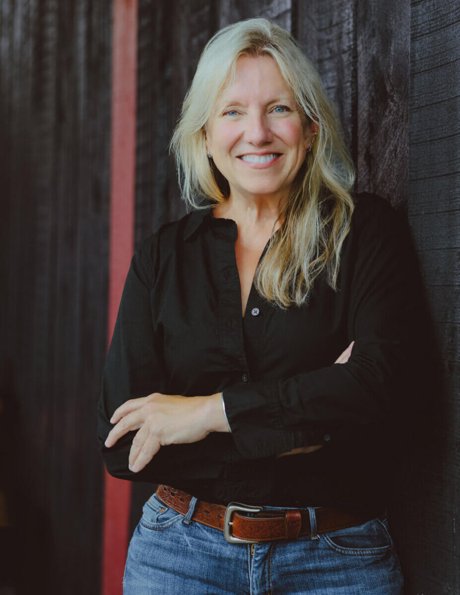 A smiling woman with blonde hair wearing a black shirt and jeans, standing with her arms crossed.