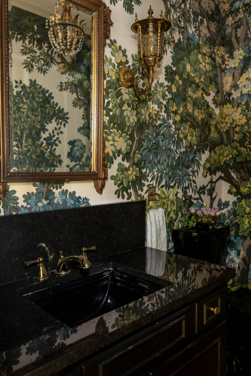 From ICU Nurse to Interior Designer: Meet Gretchen Rose of Kentucky Rose Designs - 5 A bathroom vanity with a black stone countertop and sink, featuring a gold-framed mirror, a brass wall sconce, and botanical tree wallpaper in a design by Kentucky Rose Designs.