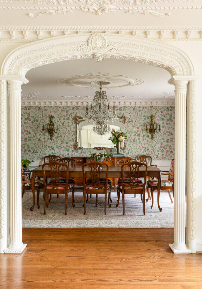 From ICU Nurse to Interior Designer: Meet Gretchen Rose of Kentucky Rose Designs - 8 Dining room seen through an arched doorway, featuring a wooden table with six chairs, floral wallpaper, a crystal chandelier, and an ornate mirror in a style by interior designer Gretchen Rose.