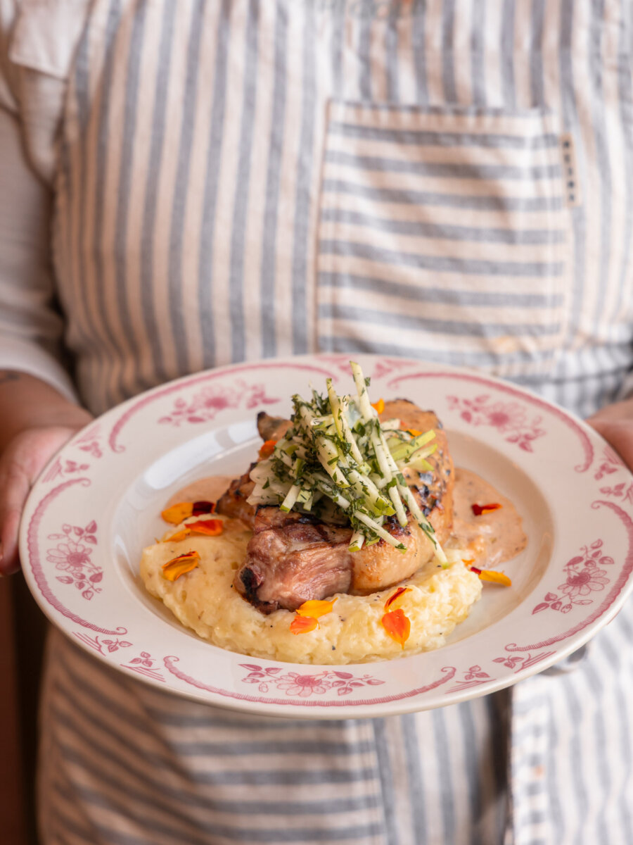 A person in a striped apron holds a plate of pork chops and mashed potatoes topped with shredded apple and flower petals, a highlight of new Louisville restaurants.