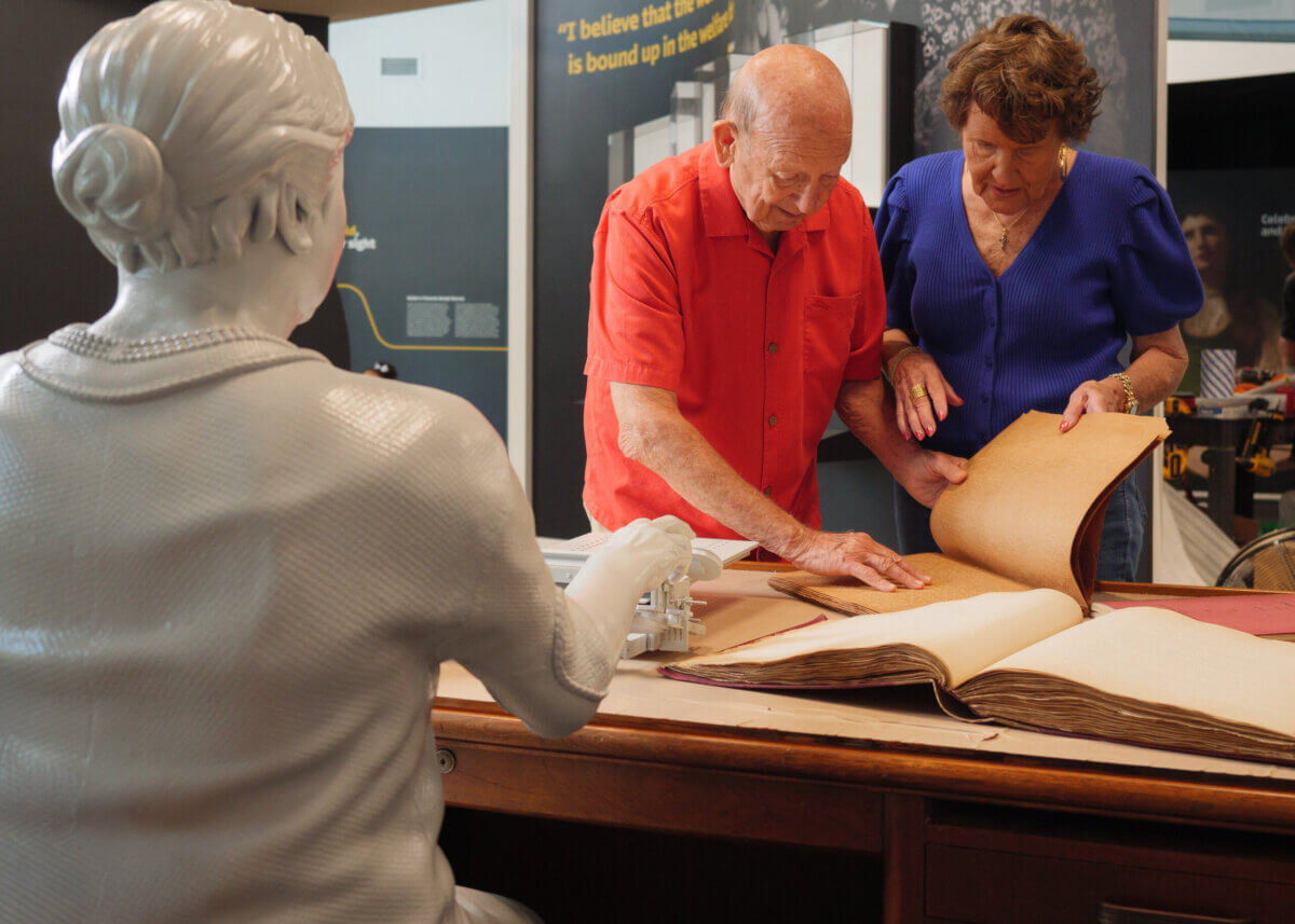Stevie Wonder's Piano, Helen Keller's Desk & the Louisville Museum Where Every Guest Gets to Touch History - 8 An elderly couple looks through a large, open book at The Dot Experience, sitting at a table next to a statue of a woman at a desk.