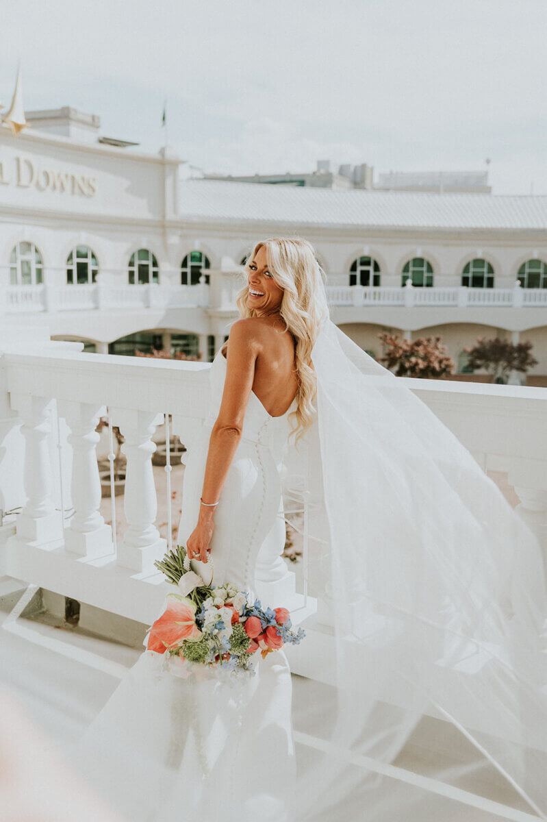 Bride in a white dress holding a colorful bouquet stands on a balcony at the Kentucky Derby Museum, smiling over her shoulder with her veil flowing in the wind.
