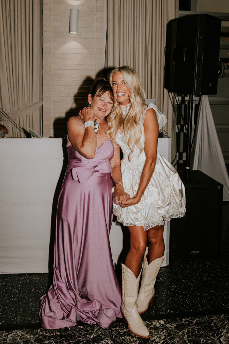 Two women pose together indoors at a wedding reception; one in a lavender gown, the other in a short white dress with cowboy boots—something old, new, and blue.