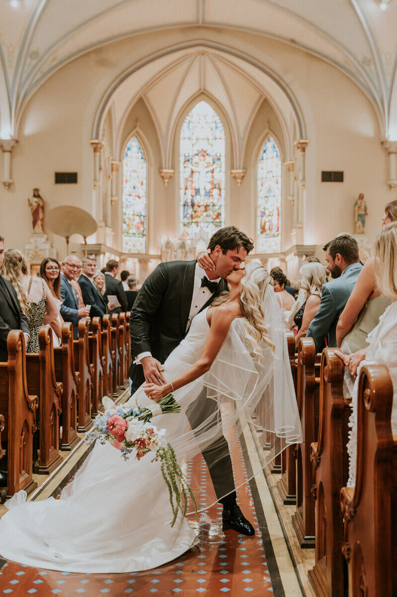 A bride and groom kiss in the aisle of a church after their wedding, ready to celebrate at their wedding reception surrounded by guests and stained glass windows.