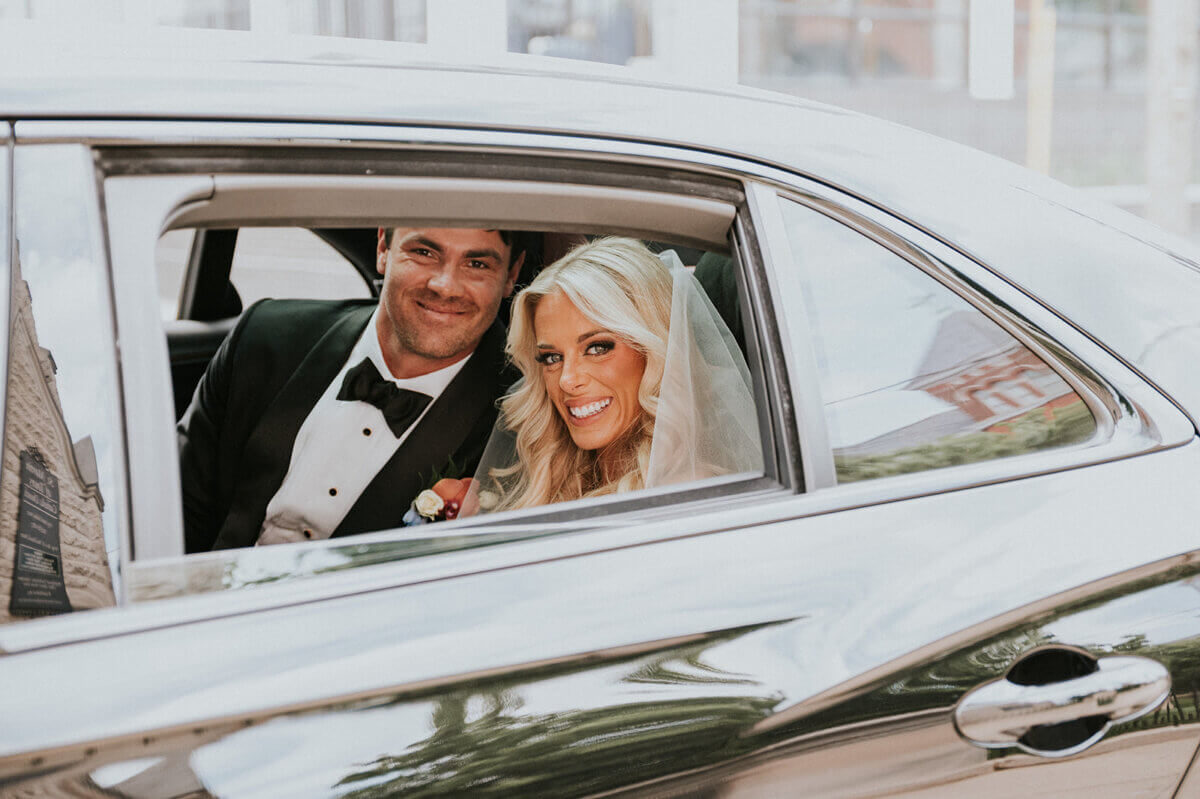 A bride and groom in wedding attire smile while sitting in the back seat of a car, embracing the spirit of something old something new as they head to their wedding reception.