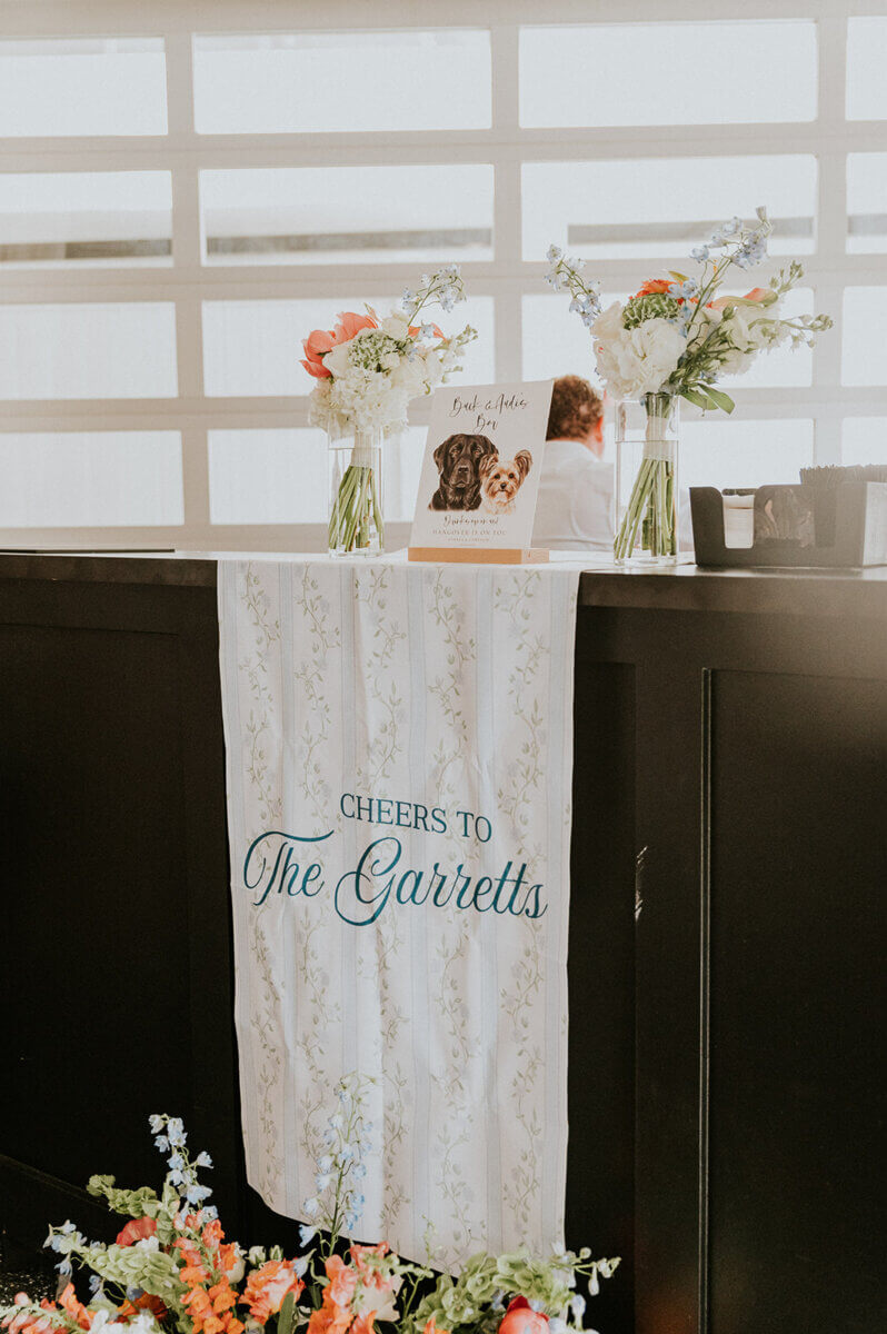 A bar counter with a sign reading “Cheers to The Garretts,” two vases of flowers, and a framed photo of a dog at this elegant wedding reception.