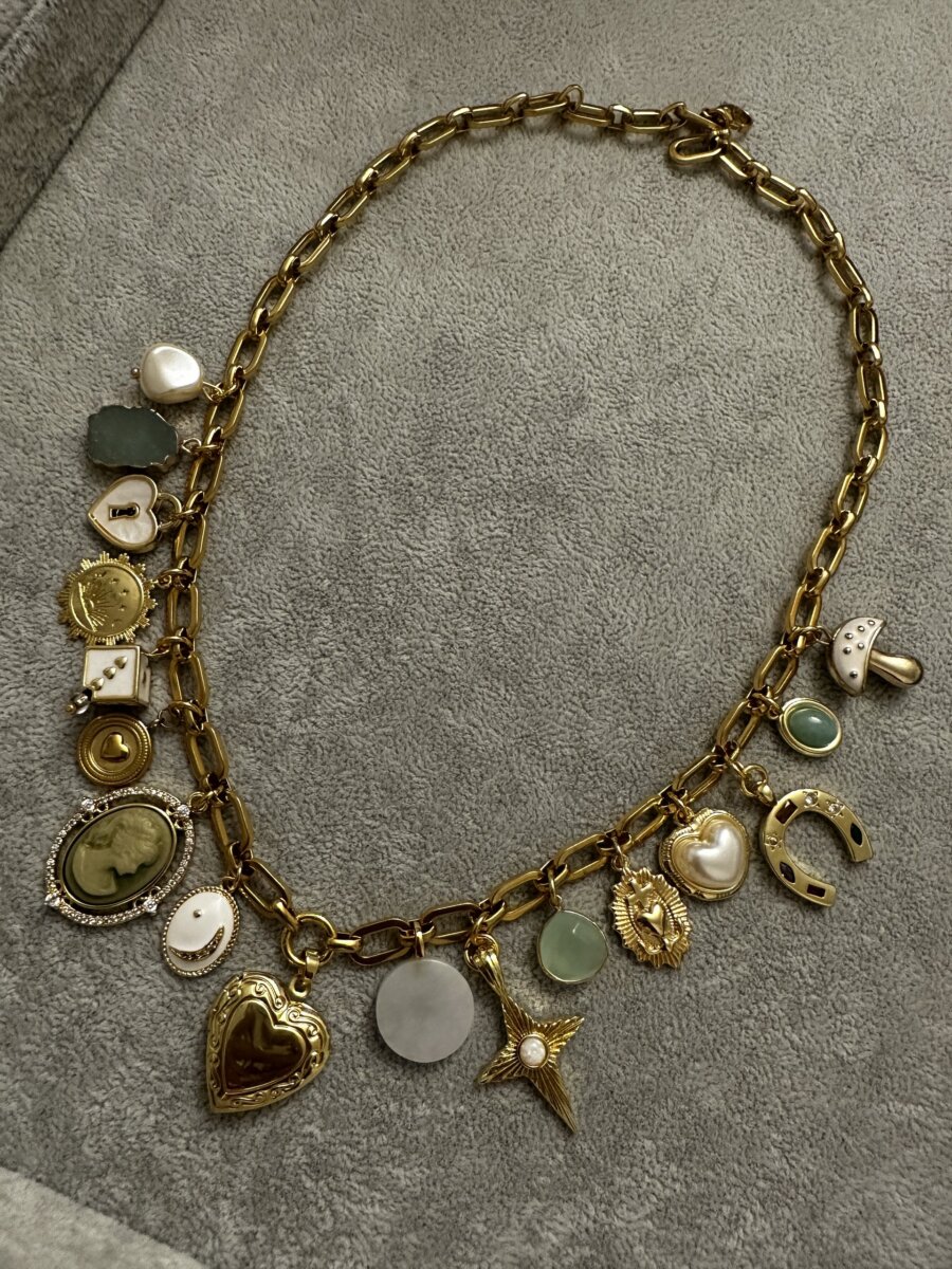 Gold chain charm necklace featuring assorted charms including hearts, a cameo, a horseshoe, a star, and a mushroom on a gray surface.