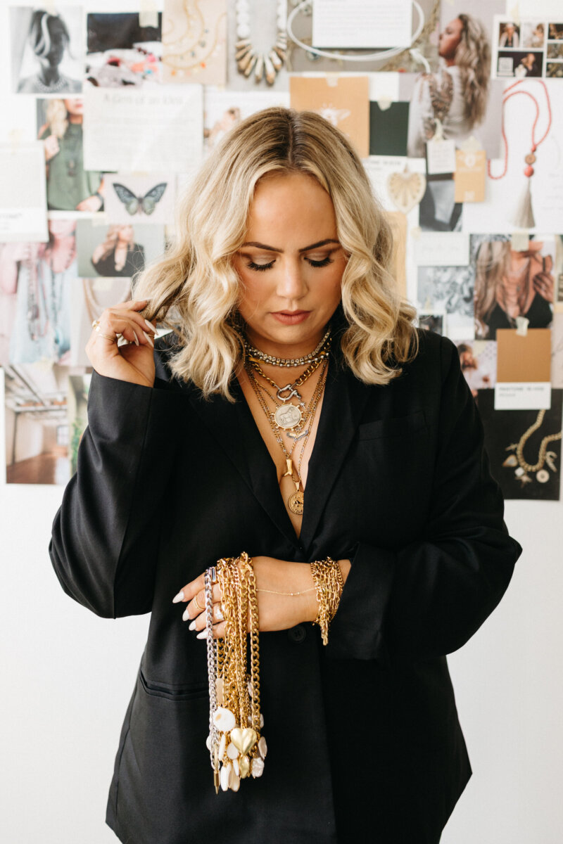 A woman wearing a black blazer and multiple layered gold necklaces holds a collection of gold chain bracelets in her hand.