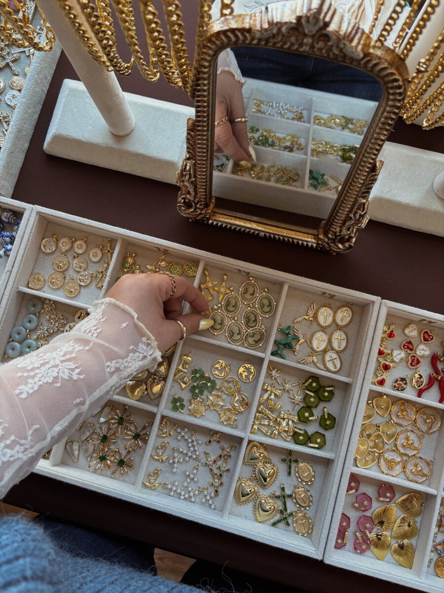 A hand reaching into a jewelry display case filled with small gold-toned charms, reflecting in a vintage-style mirror.