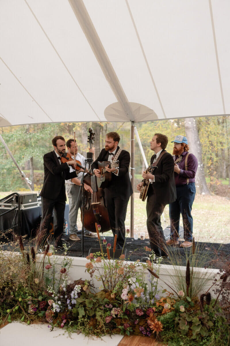 Forget Trends! Your Wedding Florals Should Show What You Love - 6 A five-piece band with a fiddle, upright bass, guitar, and banjo plays under a white tent decorated by Snapdragon Floral.