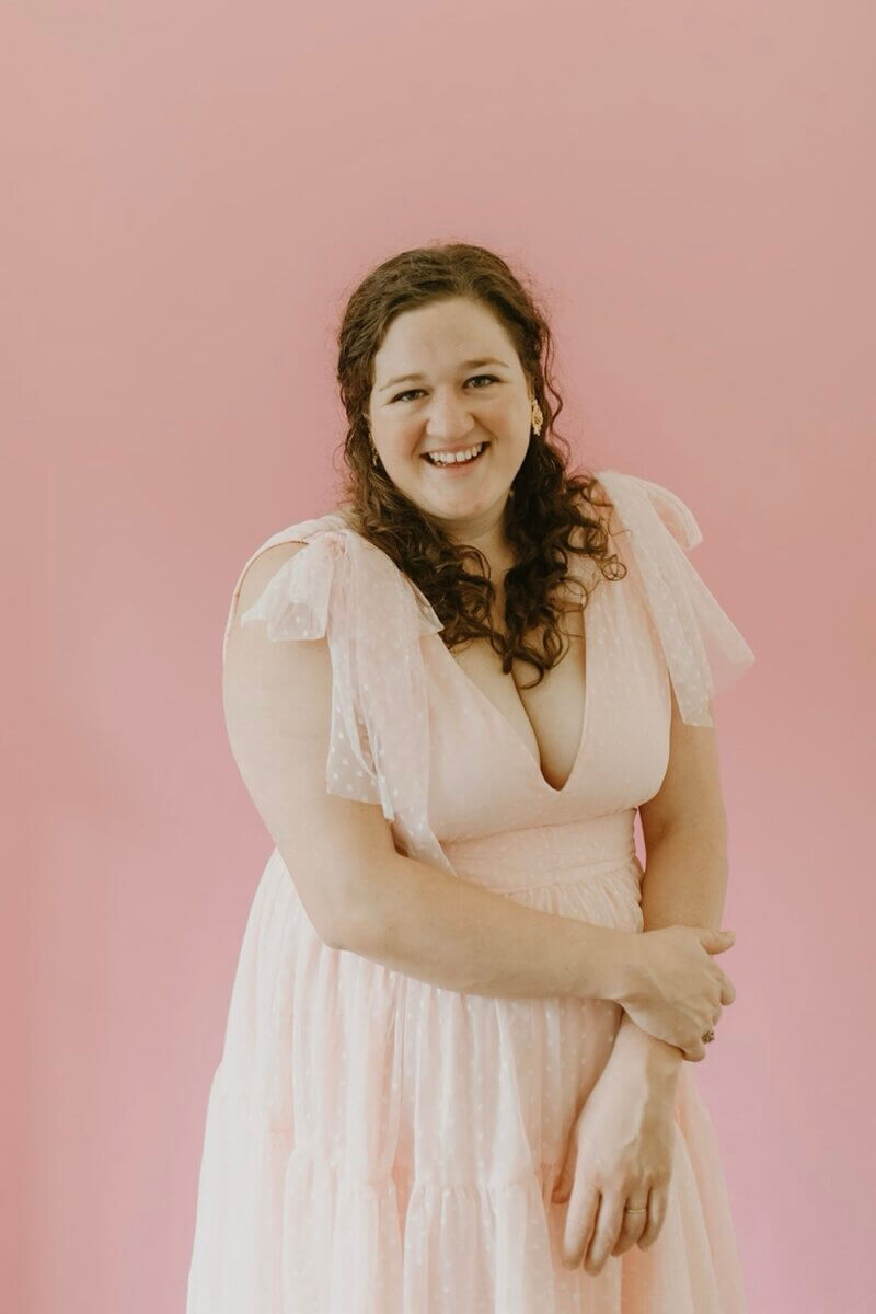 Forget Trends! Your Wedding Florals Should Show What You Love - 2 A smiling woman wearing a pale pink dress with ruffled sleeves against a solid pink background.