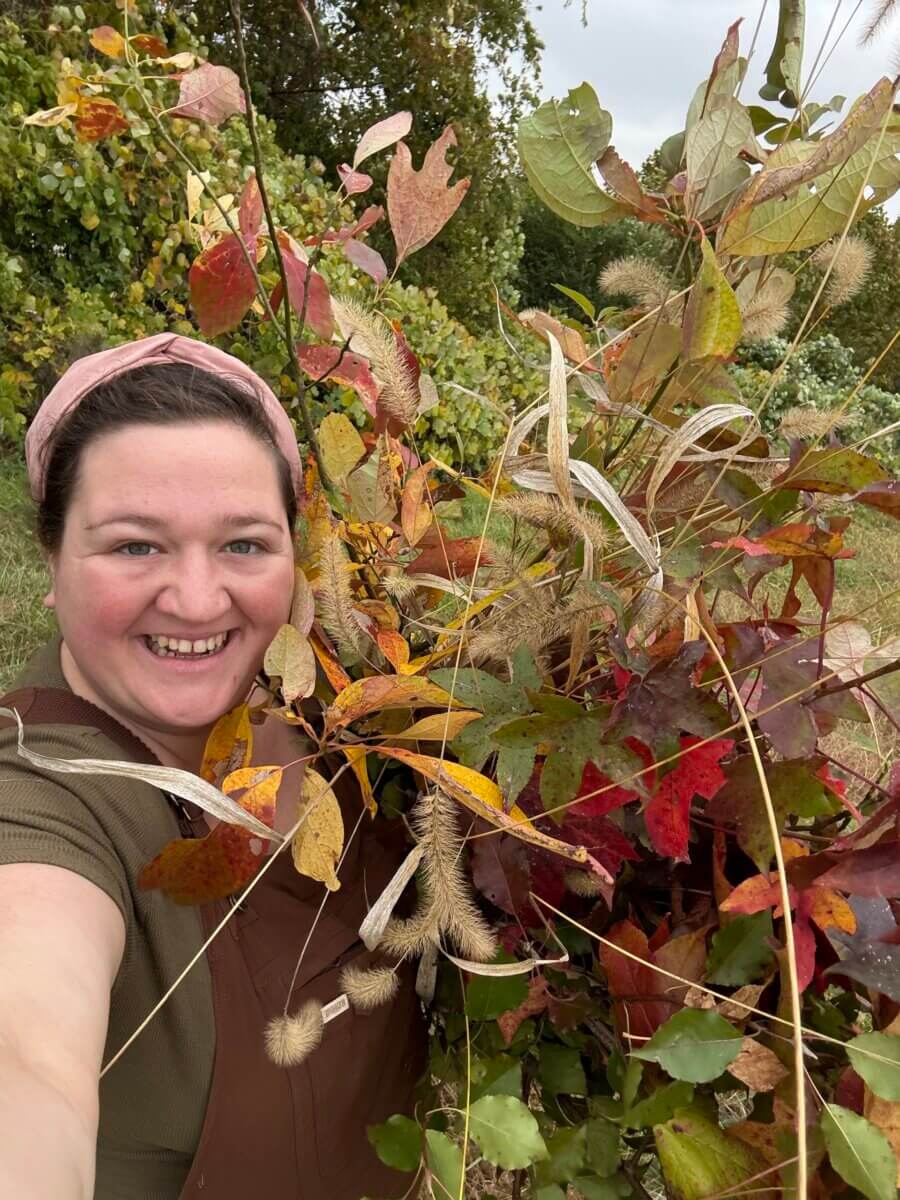 Forget Trends! Your Wedding Florals Should Show What You Love - 4 A Memphis florist wearing a pink headband and brown overalls smiles while holding a large bouquet of autumn leaves and ornamental grasses.