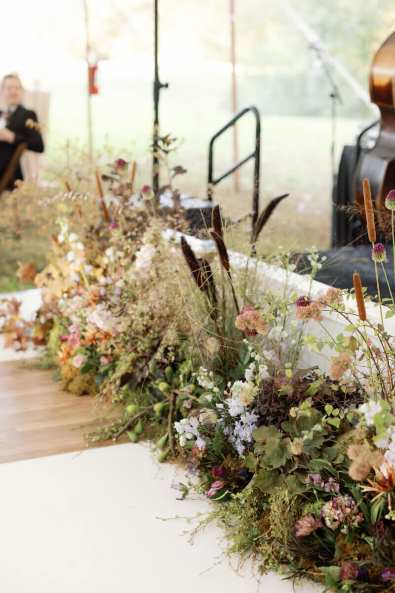Forget Trends! Your Wedding Florals Should Show What You Love - 5 A low floral arrangement of wildflowers, grasses, and cattails lining the front of a wooden stage.
