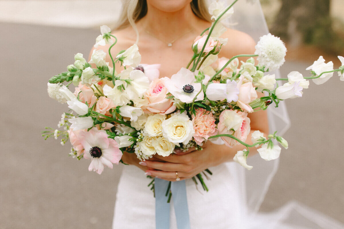 Forget Trends! Your Wedding Florals Should Show What You Love - 3 A bride in a white gown holds a bouquet of roses and anemones by Snapdragon Floral, tied with a blue ribbon.