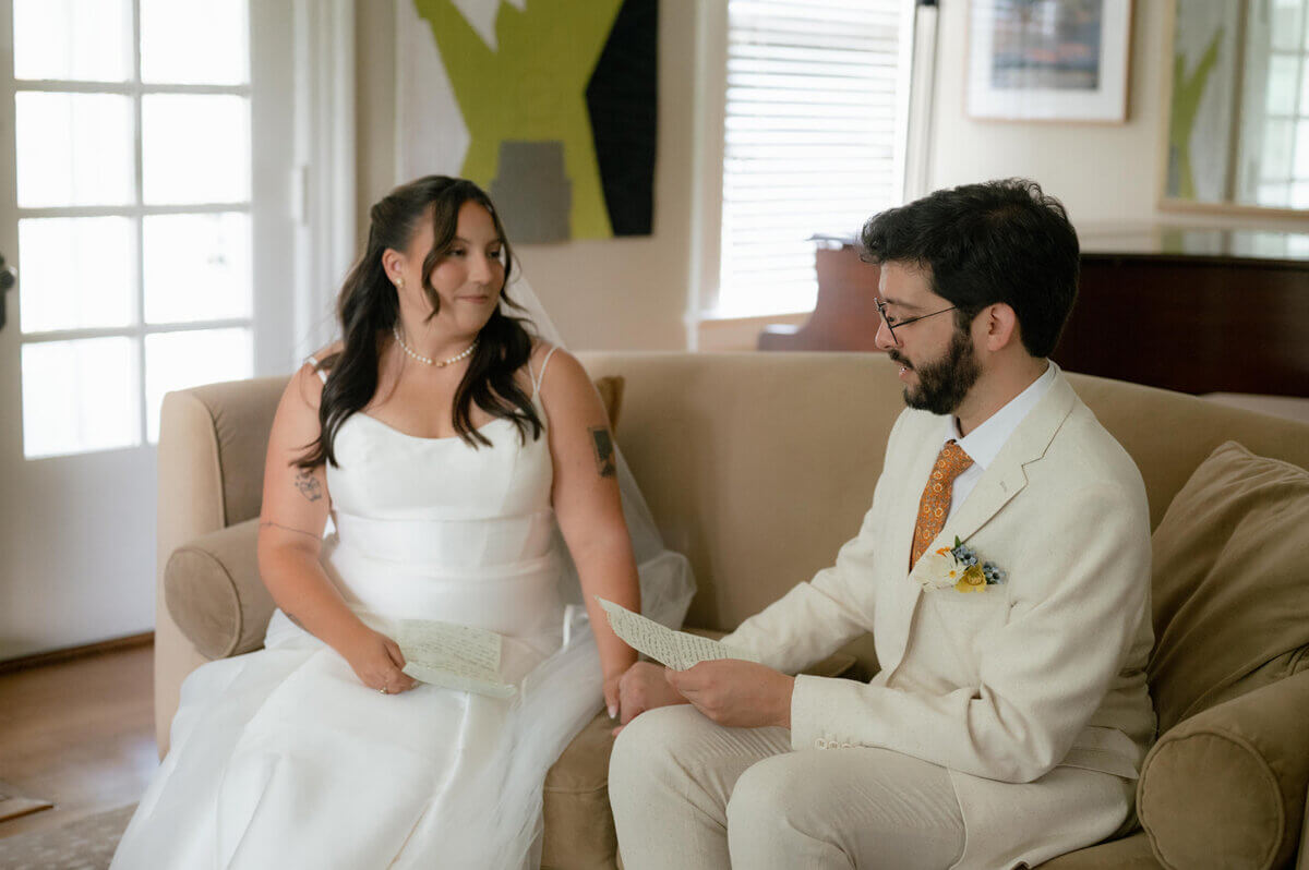This Music-centric Memphis Wedding Even Had a Tattoo Artist! - 6 A bride in a white wedding dress and a groom in a cream suit sit on a sofa reading handwritten notes to each other during their Memphis wedding.