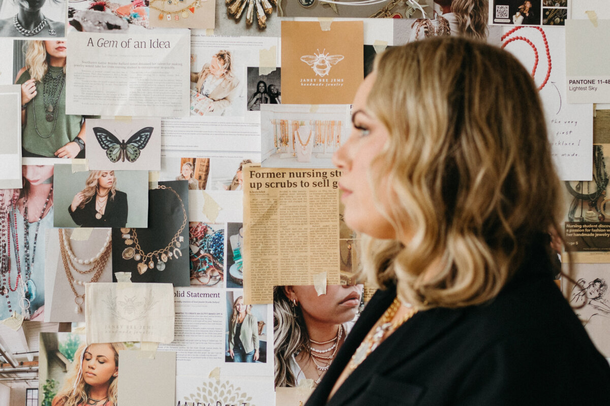A woman stands in profile before a mood board covered in photographs of Janey Bee Jems jewelry and news clippings about the business.