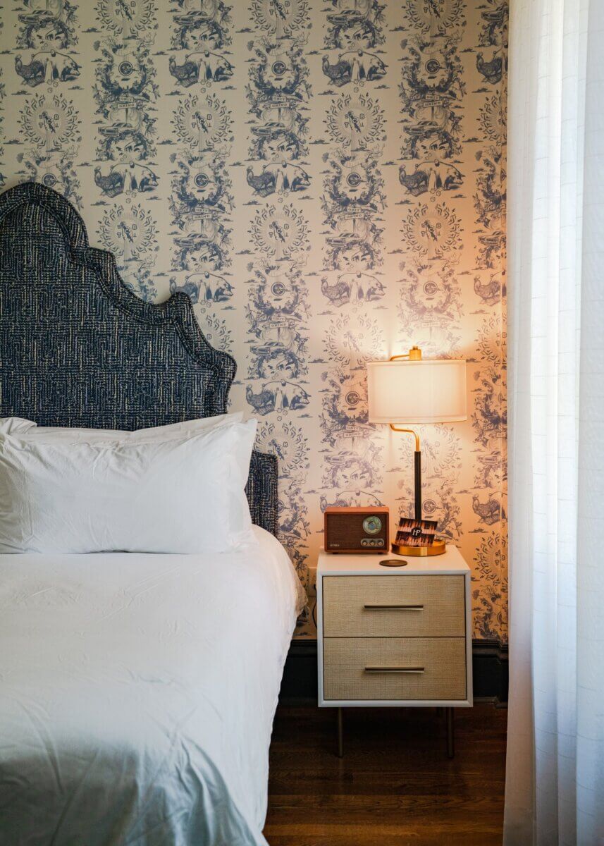 You Won't Believe What They Found Inside This 120-Year-Old Memphis Hotel - 12 A bedroom with a blue patterned headboard, a bedside table with a lamp and a vintage-style radio, against a blue and white patterned wallpaper in a boutique hotel design.