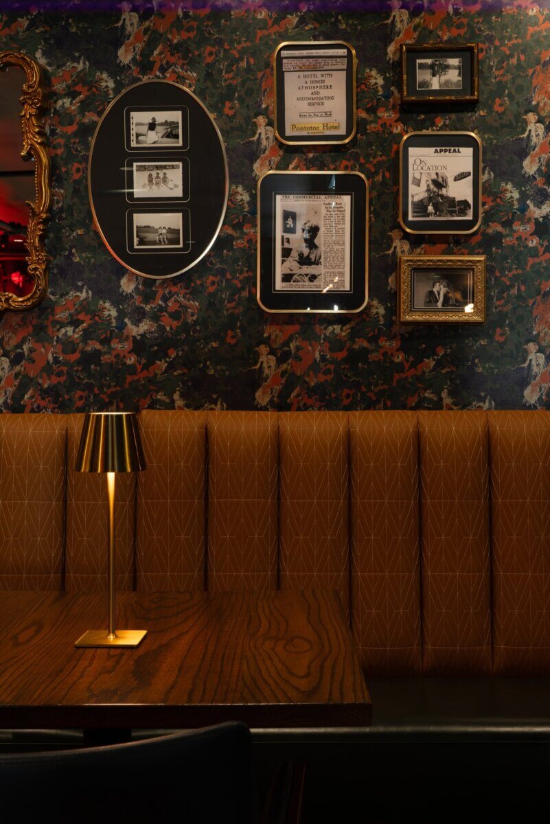 You Won't Believe What They Found Inside This 120-Year-Old Memphis Hotel - 10 A boutique hotel booth with a brown tufted bench and wooden table, illuminated by a gold lamp against floral wallpaper decorated with framed vintage advertisements and photographs.