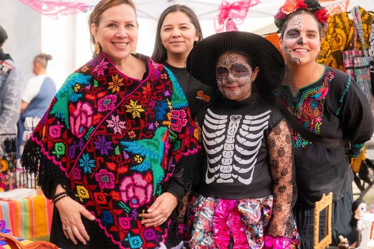 A Memphis Día de los Muertos Guide: Where to Celebrate - 3 Three women and a girl in Día de los Muertos face paint and costumes pose at a market stall featuring embroidered textiles.
