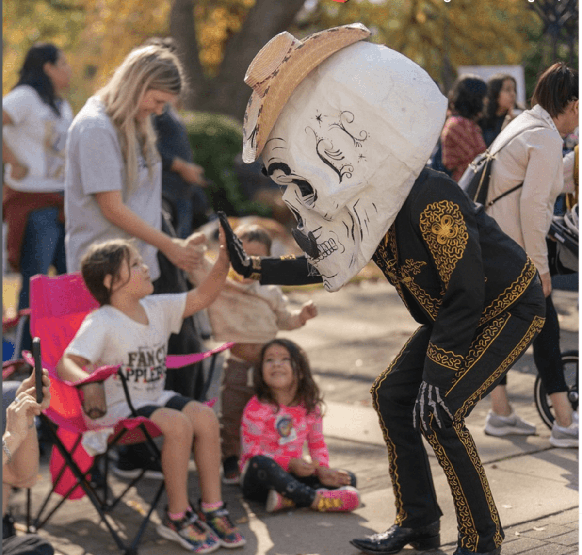 A Memphis Día de los Muertos Guide: Where to Celebrate - 4 A performer wearing a large skull mask and a charro suit high-fives a child sitting in a pink chair at an outdoor event.