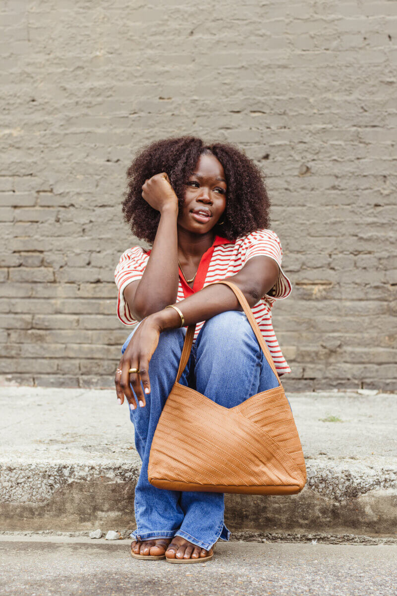 What If Your Holiday Shopping Could Change Someone's Life? - 2 A woman wearing a striped top and jeans sits on a curb, holding a tan shoulder bag.