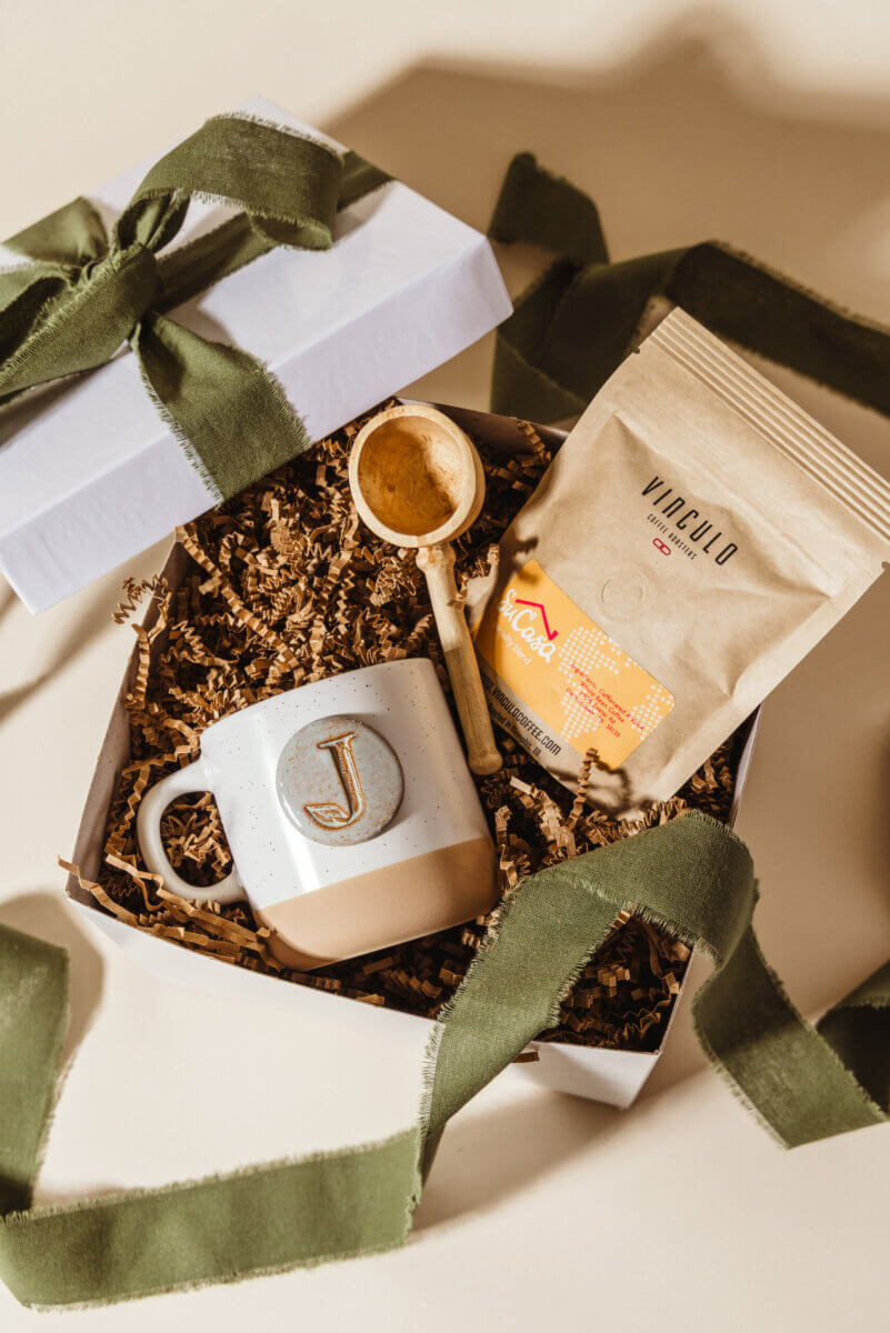What If Your Holiday Shopping Could Change Someone's Life? - 8 Gift box containing a ceramic mug with the letter J, a bag of Vinculo coffee, and a wooden scoop, decorated with a green ribbon.