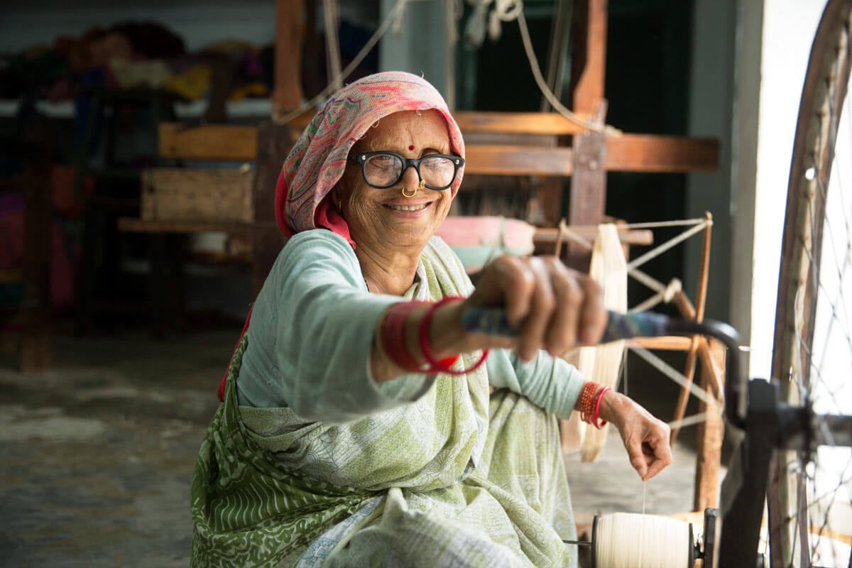 What If Your Holiday Shopping Could Change Someone's Life? - 5 An elderly woman wearing a patterned headscarf and glasses smiles while operating a spinning wheel.