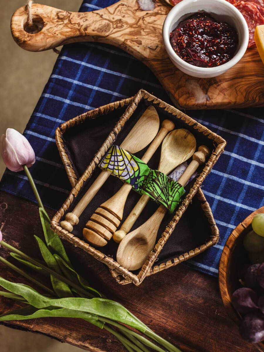 What If Your Holiday Shopping Could Change Someone's Life? - 9 A small woven basket containing three wooden serving utensils held together by a patterned fabric band, sitting on a blue checkered cloth.