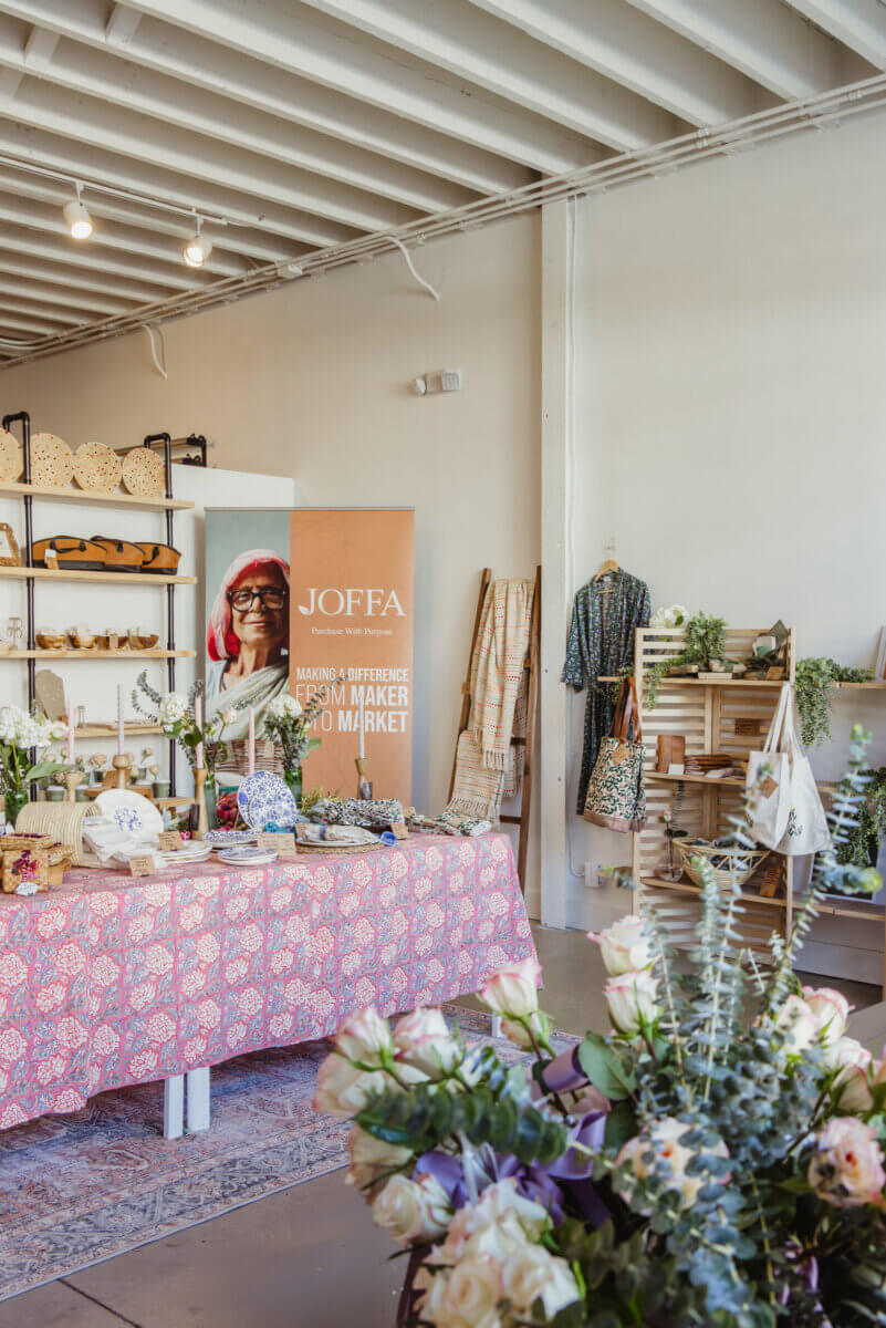 What If Your Holiday Shopping Could Change Someone's Life? - 6 A market stall featuring a JOFFA banner, a table covered in a pink floral cloth displaying ceramics and textiles, and wooden shelving with bags and decor.