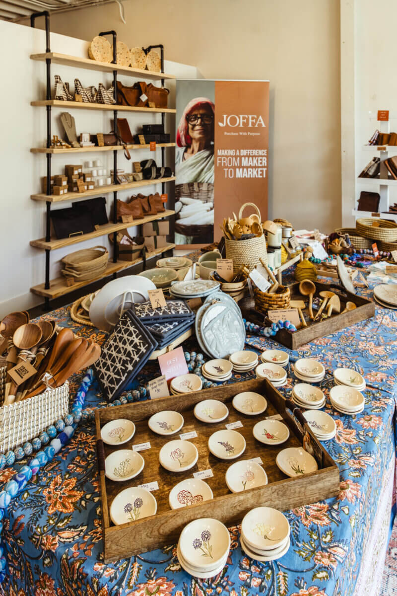 What If Your Holiday Shopping Could Change Someone's Life? - 3 A JOFFA banner stands behind tables filled with artisan goods, including wooden spoons, patterned napkins, and ceramic dishes painted with flowers.
