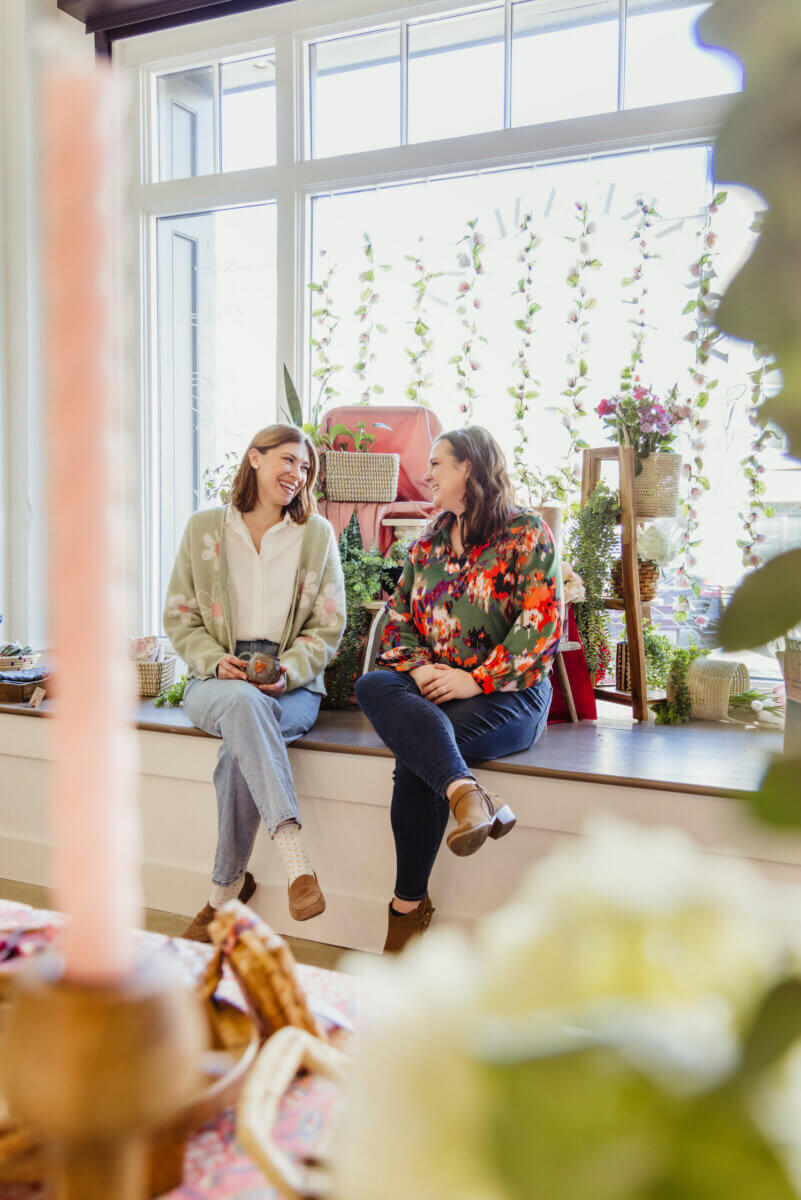 What If Your Holiday Shopping Could Change Someone's Life? - 4 Two women sitting on a window ledge, laughing and talking to each other in a shop filled with plants and decor.