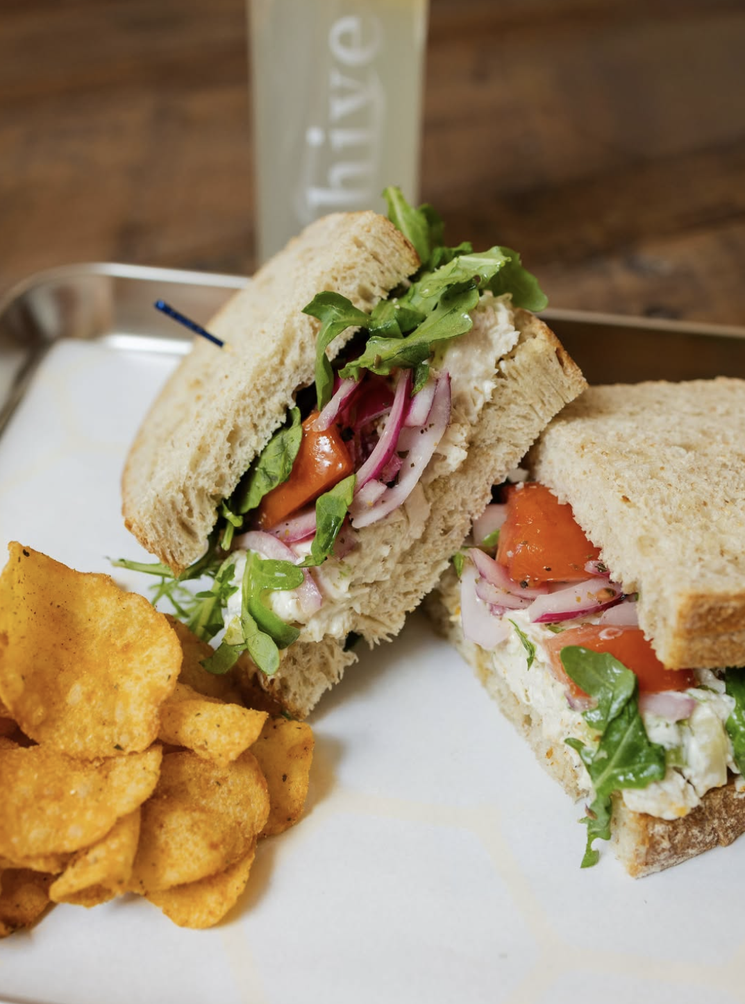 A chicken salad sandwich on wheat bread with arugula, tomato, and red onion, served with a side of potato chips.