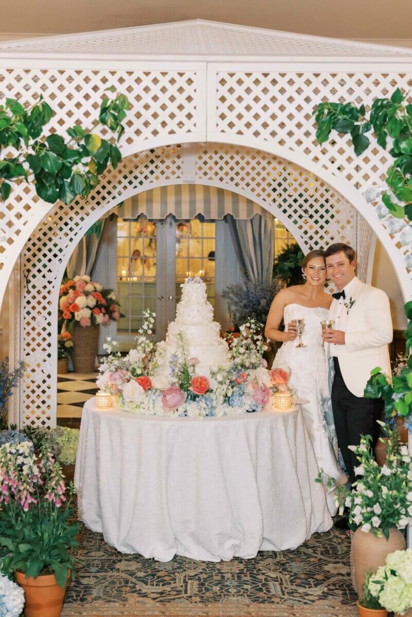 A Secret Speakeasy and Late-Night Taco Bell?! This Nashville Wedding Had Serious Surprises - 14 A bride and groom stand beside a multi-tiered wedding cake on a table adorned with garden-party florals under a white lattice archway.