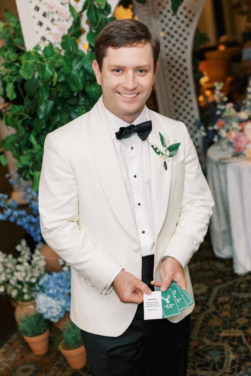 A Secret Speakeasy and Late-Night Taco Bell?! This Nashville Wedding Had Serious Surprises - 16 A man in a white tuxedo jacket and black bowtie holds a small white card and a stack of green playing cards at a Nashville garden party wedding.